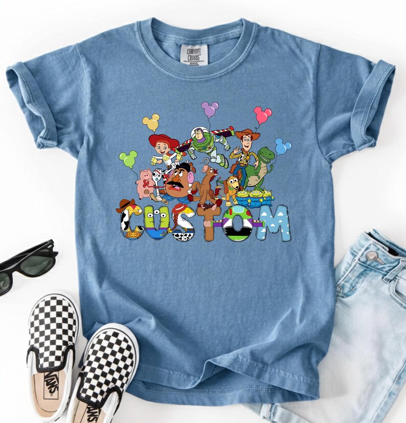 A blue t-shirt with a colorful graphic of characters from the Toy Story franchise, including Woody, Buzz Lightyear, and other beloved characters, along with the word "CUSTOM" written on it.