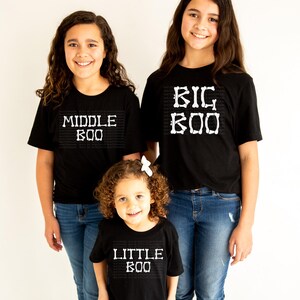 Three young girls wearing black t-shirts with the text "MIDDLE BOO" and "BIG BOO" printed on them, standing together and smiling.