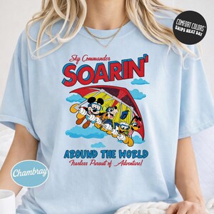 A woman wearing a light blue t-shirt with a graphic design featuring a cartoon character and the text "Sky Commander SOARIN' Around the World Fearless Pursuit of Adventures".