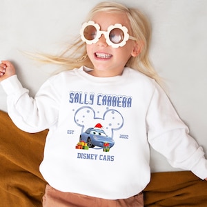 A young girl wearing a white sweater with a graphic of a blue car and Mickey Mouse ears, smiling and wearing large sunglasses.
