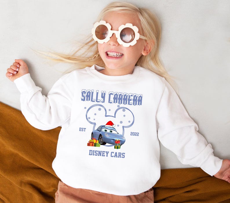 A young girl wearing a white sweater with a graphic of a blue car and Mickey Mouse ears, smiling and wearing large sunglasses.