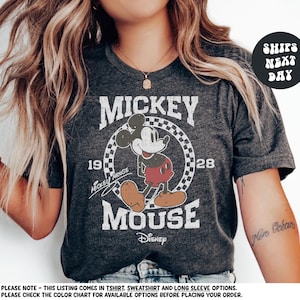 A woman wearing a gray t-shirt with a Mickey Mouse graphic and text, posing with her hair blowing in the wind.