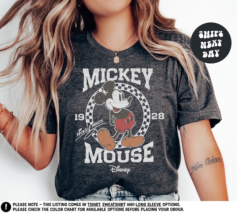 A woman wearing a gray t-shirt with a Mickey Mouse graphic and text, posing with her hair blowing in the wind.