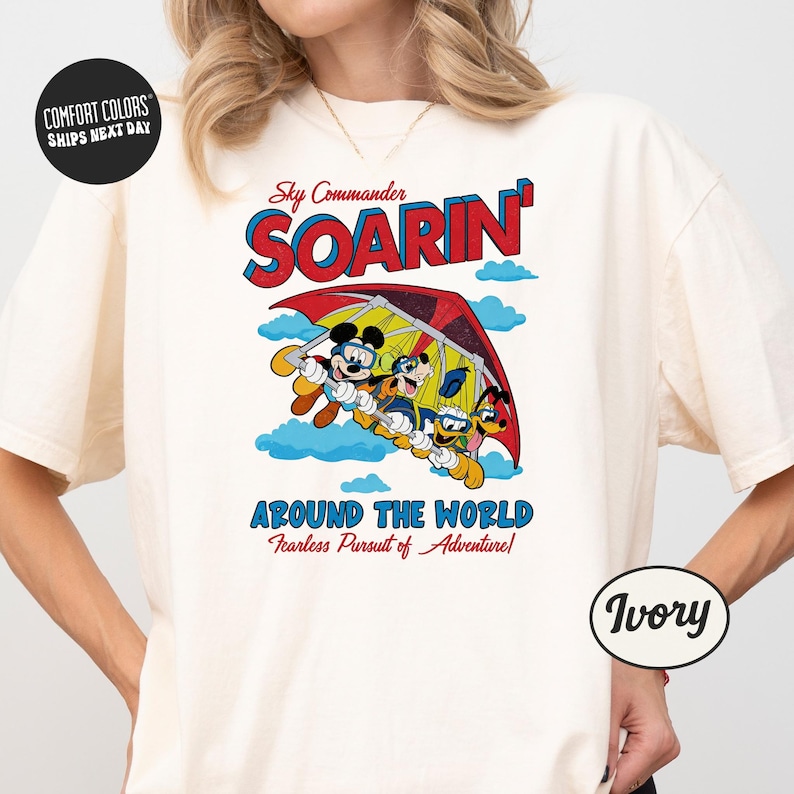 A white t-shirt with a colorful graphic design featuring a group of Disney characters, including Mickey Mouse and Donald Duck, flying through the sky.