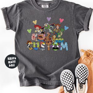 A gray t-shirt with a colorful graphic of characters from the Toy Story franchise, including Buzz Lightyear, Woody, and other beloved characters, along with the word "CUSTOM" prominently displayed.