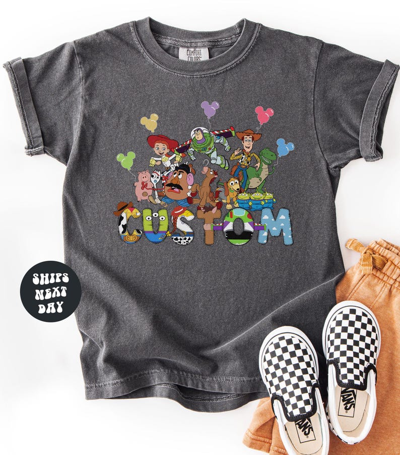 A gray t-shirt with a colorful graphic of characters from the Toy Story franchise, including Buzz Lightyear, Woody, and other beloved characters, along with the word "CUSTOM" prominently displayed.