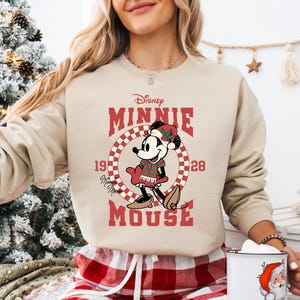 A woman wearing a beige sweater with a graphic of Minnie Mouse and the text "Disney Minnie Mouse" is holding a mug in front of a Christmas tree.