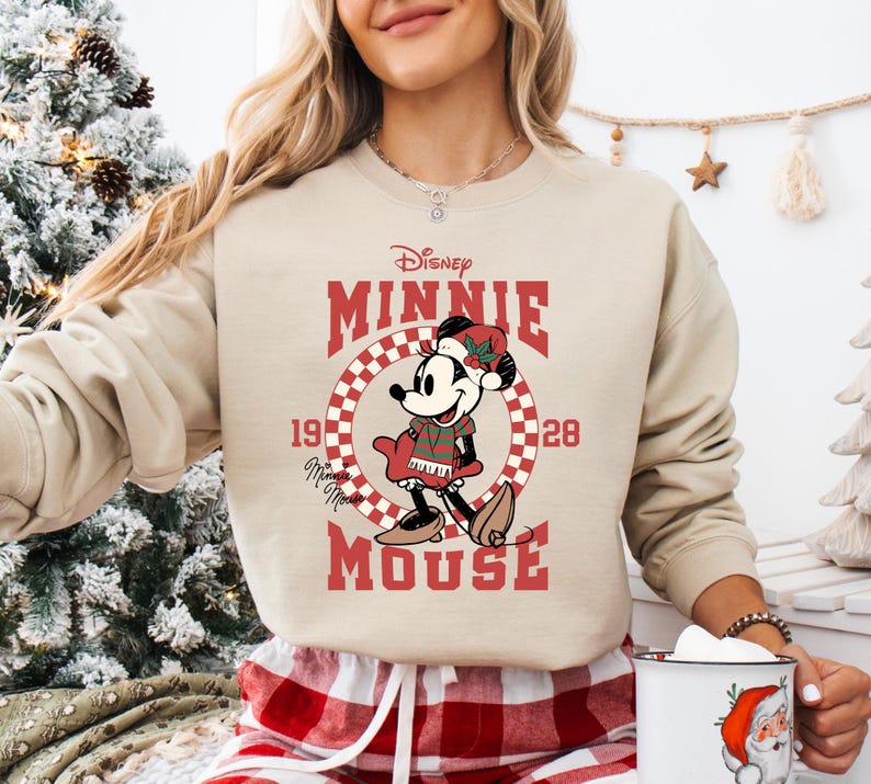 A woman wearing a beige sweater with a graphic of Minnie Mouse and the text "Disney Minnie Mouse" is holding a mug in front of a Christmas tree.