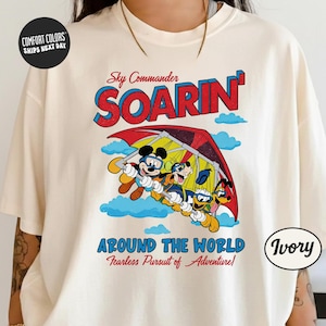 A white t-shirt with a colorful graphic design featuring Mickey Mouse and Minnie Mouse on a hang glider, along with the text "Sky Commander SOARIN' Around the World Fearless Pursuit of Adventure".