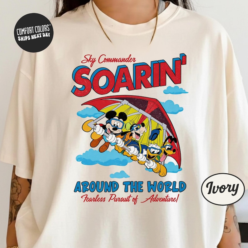 A white t-shirt with a colorful graphic design featuring Mickey Mouse and Minnie Mouse on a hang glider, along with the text "Sky Commander SOARIN' Around the World Fearless Pursuit of Adventure".
