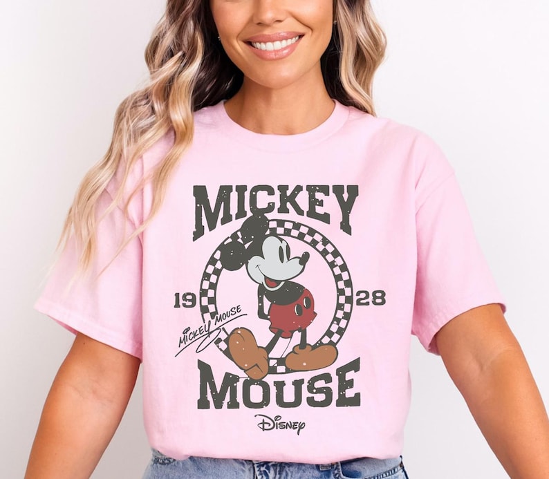 A woman wearing a pink t-shirt with a Mickey Mouse graphic and text, smiling at the camera.