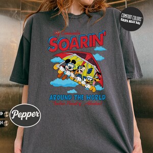 A gray t-shirt with a graphic design featuring a group of cartoon characters, including Mickey Mouse and Goofy, flying in a hot air balloon. The text on the t-shirt reads "Soarin' Around the World" and "Pepsi".