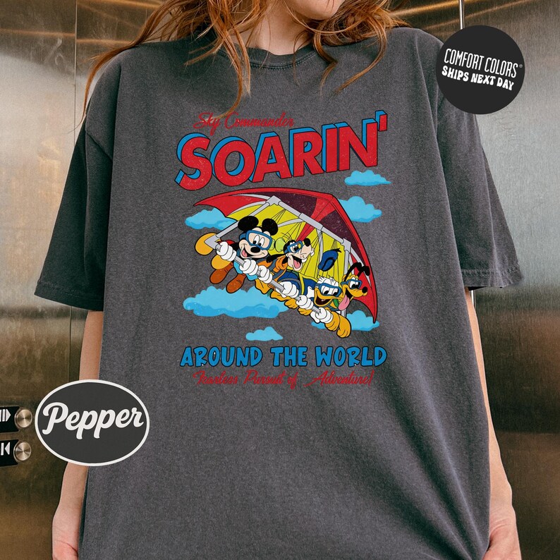 A gray t-shirt with a graphic design featuring a group of cartoon characters, including Mickey Mouse and Goofy, flying in a hot air balloon. The text on the t-shirt reads "Soarin' Around the World" and "Pepsi".