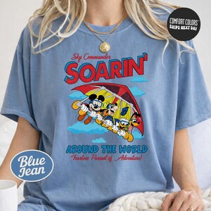A woman wearing a blue t-shirt with a graphic design featuring Mickey Mouse and Goofy, along with the text "Soarin' Around the World" and "Comfort Colors" printed on it.