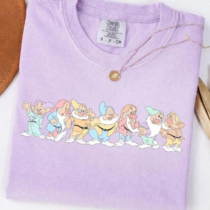 May include: A light purple t-shirt featuring a graphic of the seven dwarfs from Snow White. The shirt has a crew neck and short sleeves. A gold necklace with a pendant is draped across the shirt.