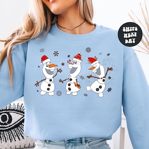 May include: Light blue sweatshirt featuring a graphic of three snowmen wearing Santa hats and surrounded by snowflakes. The snowmen have carrot noses and stick arms. The text "SHIPS NEXT DAY" is in a circle on the right side.