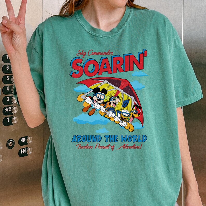 A person wearing a green t-shirt with a graphic design featuring Mickey Mouse and Goofy, along with the text "Soarin' Around the World" and "Fearless Pursuit of Adventures".