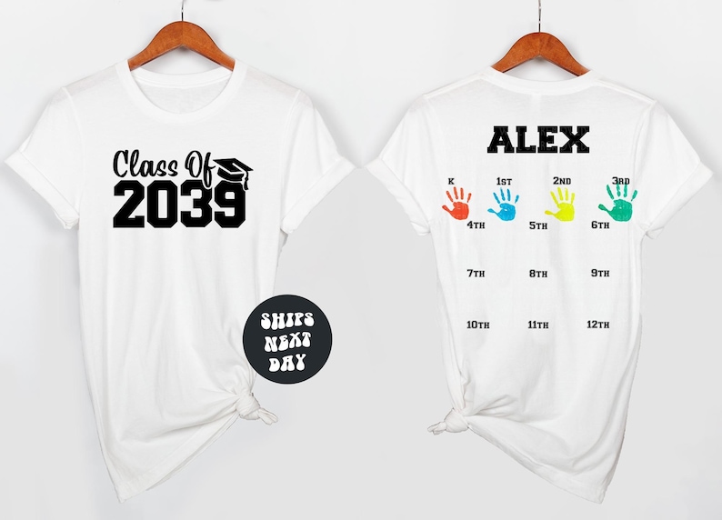 a white t-shirt with a graduation cap and mortarboard design, along with handprints representing the years from 1st to 12th. The t-shirt is displayed on a wooden hanger, and there is a text overlay that reads "Class Of 2039" and "ALEX".