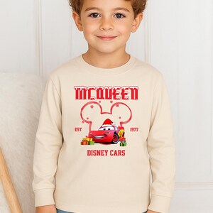 A young boy wearing a beige long-sleeved shirt with a graphic of a red car and the text "McQueen" and "Disney Cars" printed on it.