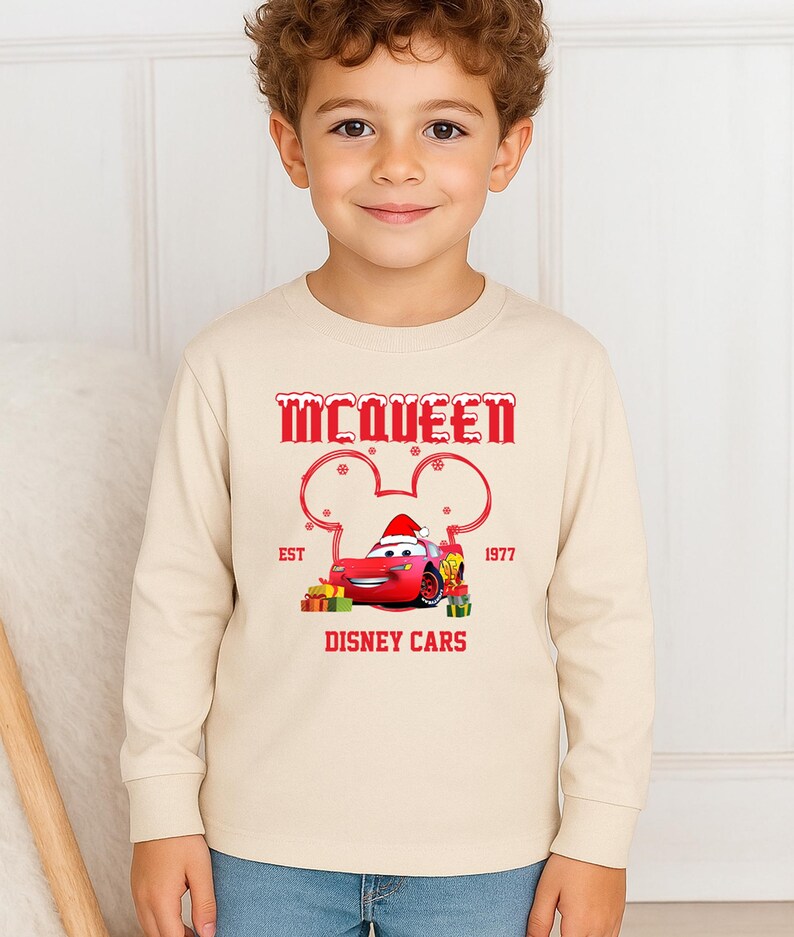 A young boy wearing a beige long-sleeved shirt with a graphic of a red car and the text "McQueen" and "Disney Cars" printed on it.