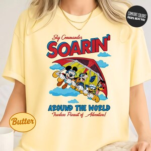 A yellow t-shirt with a graphic design featuring a cartoon character and the text "Soarin' Around the World" and "Fearless Pursuit of Adventure".
