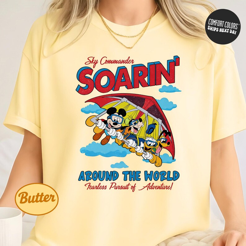 A yellow t-shirt with a graphic design featuring a cartoon character and the text "Soarin' Around the World" and "Fearless Pursuit of Adventure".