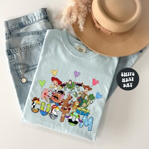 A light blue t-shirt with a colorful graphic of characters from the Toy Story movie, including Woody, Buzz Lightyear, and other beloved characters, along with a hat and some jeans.