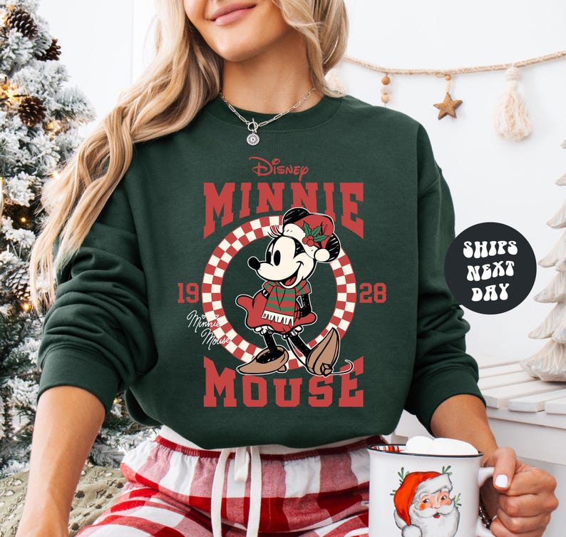 A woman wearing a green sweatshirt with a graphic of Minnie Mouse and the text "Disney MINNIE MOUSE" is holding a mug with a Santa Claus design.
