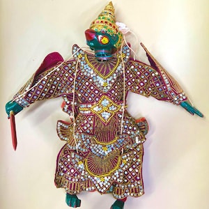 May include: A green puppet with a purple and gold costume, adorned with sequins and beads. The puppet has a crown and wings and is holding a red sword.