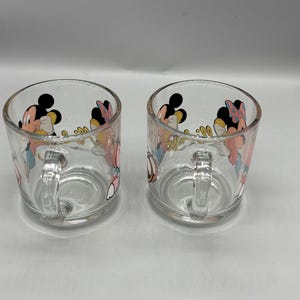 May include: Two clear glass mugs with handles. Each mug features a colorful cartoon design of Mickey and Minnie Mouse. The mugs are cylindrical with a rounded base and a small handle on the side. The cartoon characters are in bright colors.