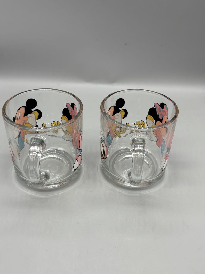 May include: Two clear glass mugs with handles. Each mug features a colorful cartoon design of Mickey and Minnie Mouse. The mugs are cylindrical with a rounded base and a small handle on the side. The cartoon characters are in bright colors.