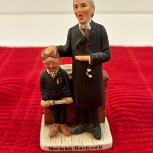 May include: A porcelain figurine set depicting a man in a black suit and a boy in a black suit holding a scroll. The man is speaking and the boy is looking up at him. The figurine is on a white base with the words "Norman Rockwell" inscribed on it.
