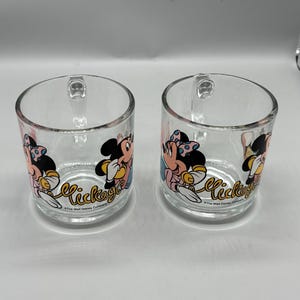 May include: Two clear glass mugs with handles. Each mug features a colorful graphic of Mickey Mouse and Minnie Mouse. The word "Mickey" is written in a cursive font on the front of each mug. The mugs are from The Walt Disney Company.