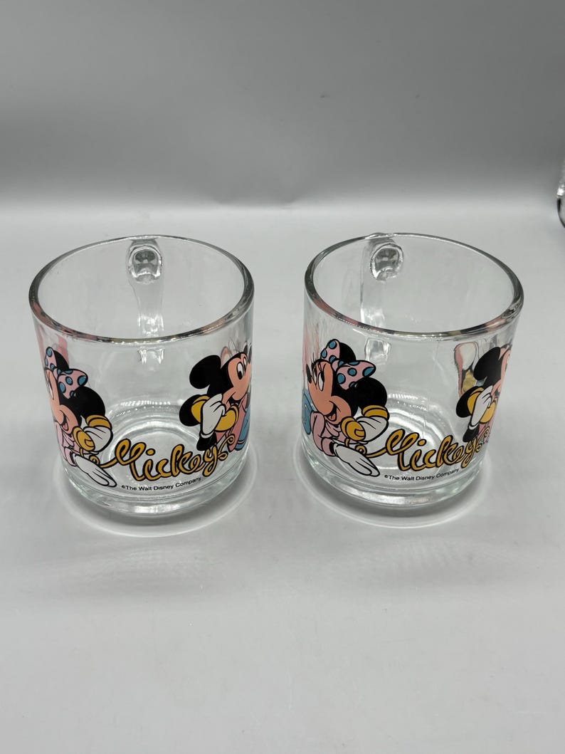 May include: Two clear glass mugs with handles. Each mug features a colorful graphic of Mickey Mouse and Minnie Mouse. The word "Mickey" is written in a cursive font on the front of each mug. The mugs are from The Walt Disney Company.