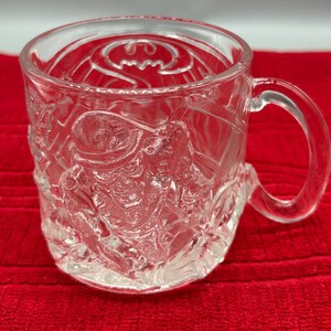 May include: Clear glass mug featuring embossed Batman imagery. The mug has a handle and a detailed design of the Batman logo on the top rim. The sides of the mug showcase Batman in action. The mug is sitting on a red surface.