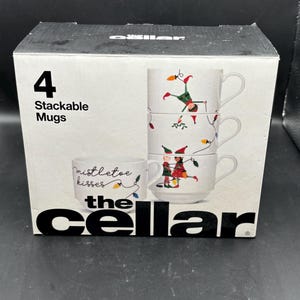 May include: A box set of four stackable white ceramic mugs. Each mug features a festive design of elves and Christmas lights. One mug has the text "mistletoe kisses". The box is black with the text "the Cellar".
