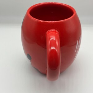 May include: A red ceramic mug with a rounded shape and a large handle. The mug features a black and white cartoon character design on the side. White text is visible on the side of the mug. The mug is designed for hot beverages.
