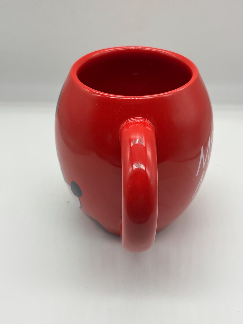 May include: A red ceramic mug with a rounded shape and a large handle. The mug features a black and white cartoon character design on the side. White text is visible on the side of the mug. The mug is designed for hot beverages.