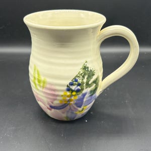 May include: A white ceramic mug with a floral design. The mug has a handle and is decorated with pink, blue, yellow, and green flowers.