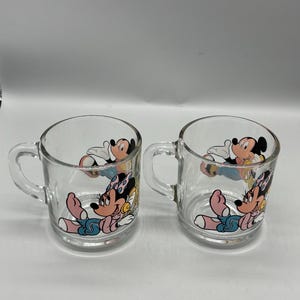 May include: Two clear glass mugs with handles. Each mug features a colorful cartoon image of Minnie Mouse and Mickey Mouse. The cartoon characters are in a relaxed pose, with Minnie Mouse wearing a pink bow.