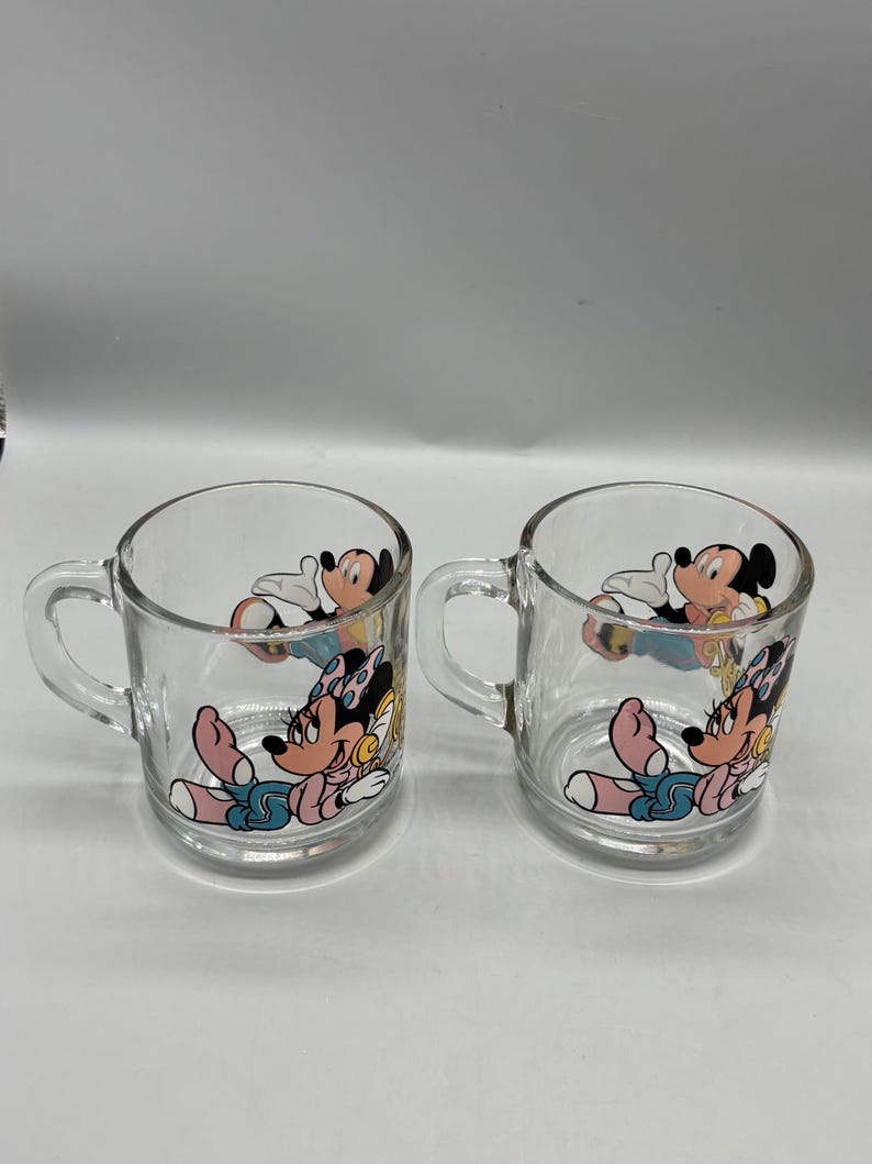 May include: Two clear glass mugs with handles. Each mug features a colorful cartoon image of Minnie Mouse and Mickey Mouse. The cartoon characters are in a relaxed pose, with Minnie Mouse wearing a pink bow.