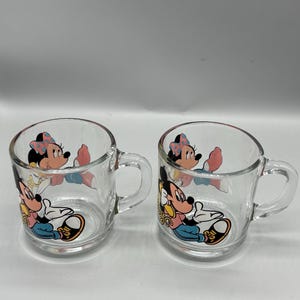 May include: Two clear glass mugs with handles. Each mug features a colorful graphic of Mickey and Minnie Mouse. The cartoon characters are depicted in a seated position. The mugs are suitable for hot or cold beverages.