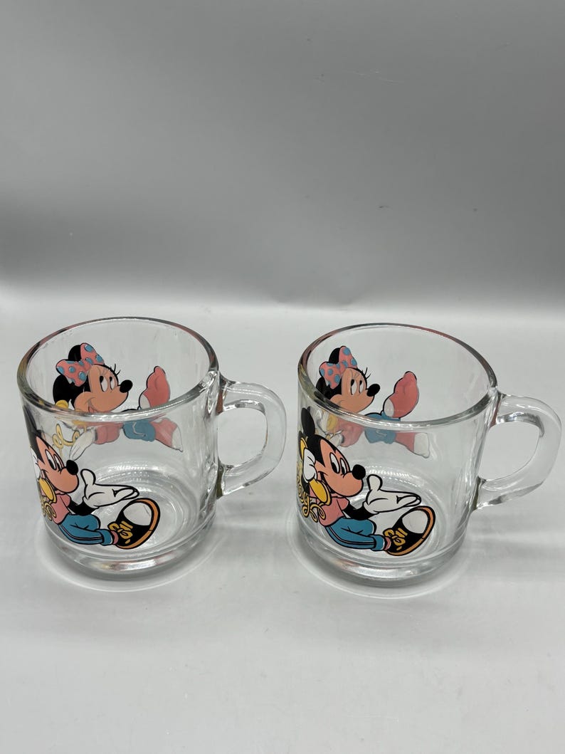 May include: Two clear glass mugs with handles. Each mug features a colorful graphic of Mickey and Minnie Mouse. The cartoon characters are depicted in a seated position. The mugs are suitable for hot or cold beverages.