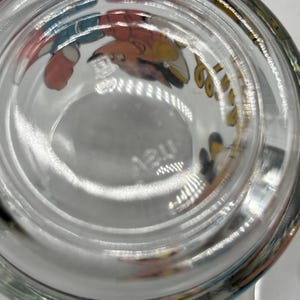 May include: Clear glass drinking glass with cartoon character illustrations around the rim. The bottom of the glass has a circular indentation and the word "may" etched into the glass.