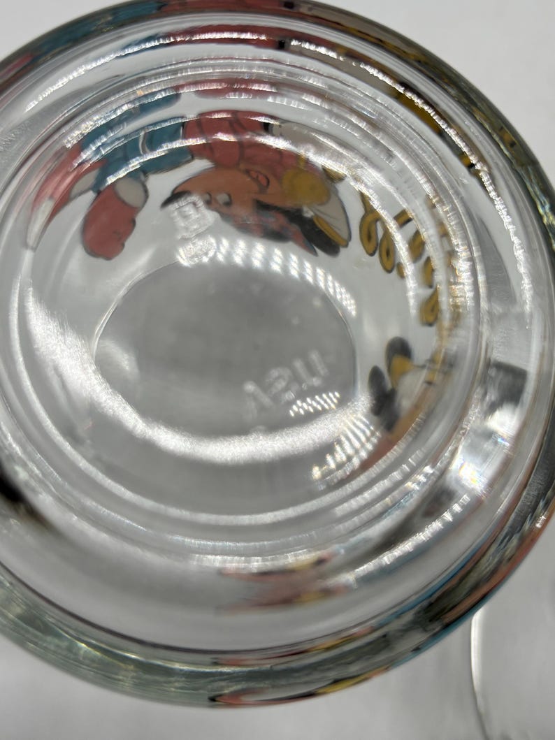 May include: Clear glass drinking glass with cartoon character illustrations around the rim. The bottom of the glass has a circular indentation and the word "may" etched into the glass.