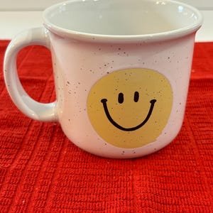 May include: White ceramic mug with a speckled finish and a yellow smiley face design.