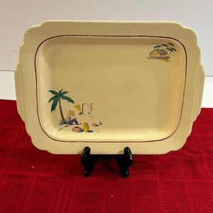 May include: A vintage yellow ceramic serving tray with a red rim and a hand-painted design of a palm tree, a person sitting under the tree, and a small house with palm trees in the background.