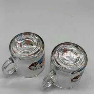 May include: Two clear glass mugs with handles, each featuring a cartoon character design. The mugs are transparent, allowing visibility of the contents. The cartoon design includes shades of blue, red, and yellow.