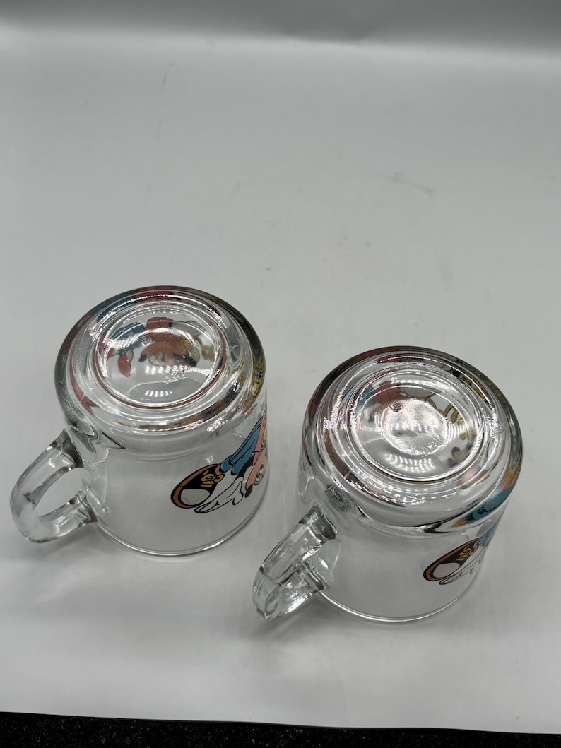 May include: Two clear glass mugs with handles, each featuring a cartoon character design. The mugs are transparent, allowing visibility of the contents. The cartoon design includes shades of blue, red, and yellow.