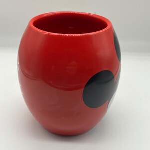 May include: A red ceramic mug with a rounded shape and a wide opening. The mug features three black circles on the side, and a white design. The mug is likely for beverages.